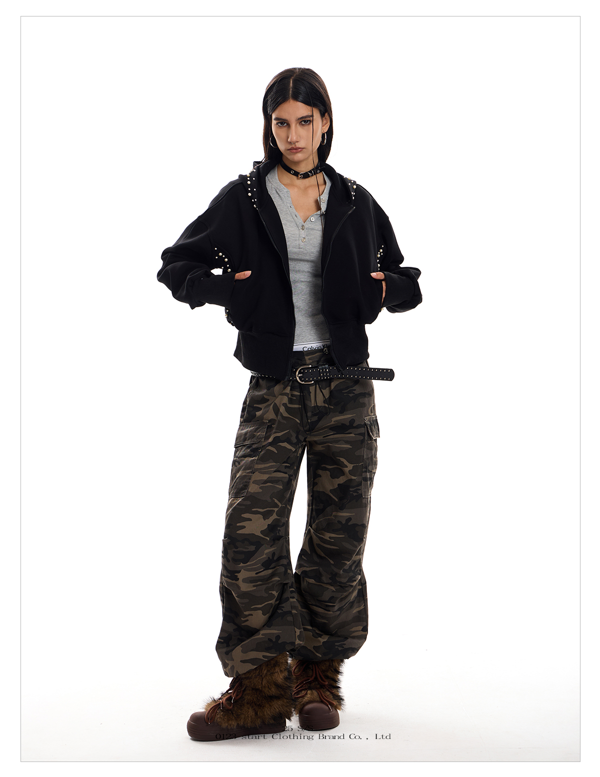 A woman models a black Street Wolf Hoodie with silver stud detailing on the sleeves and hood. She is also wearing a gray shirt, camouflage pants, and furry brown boots.