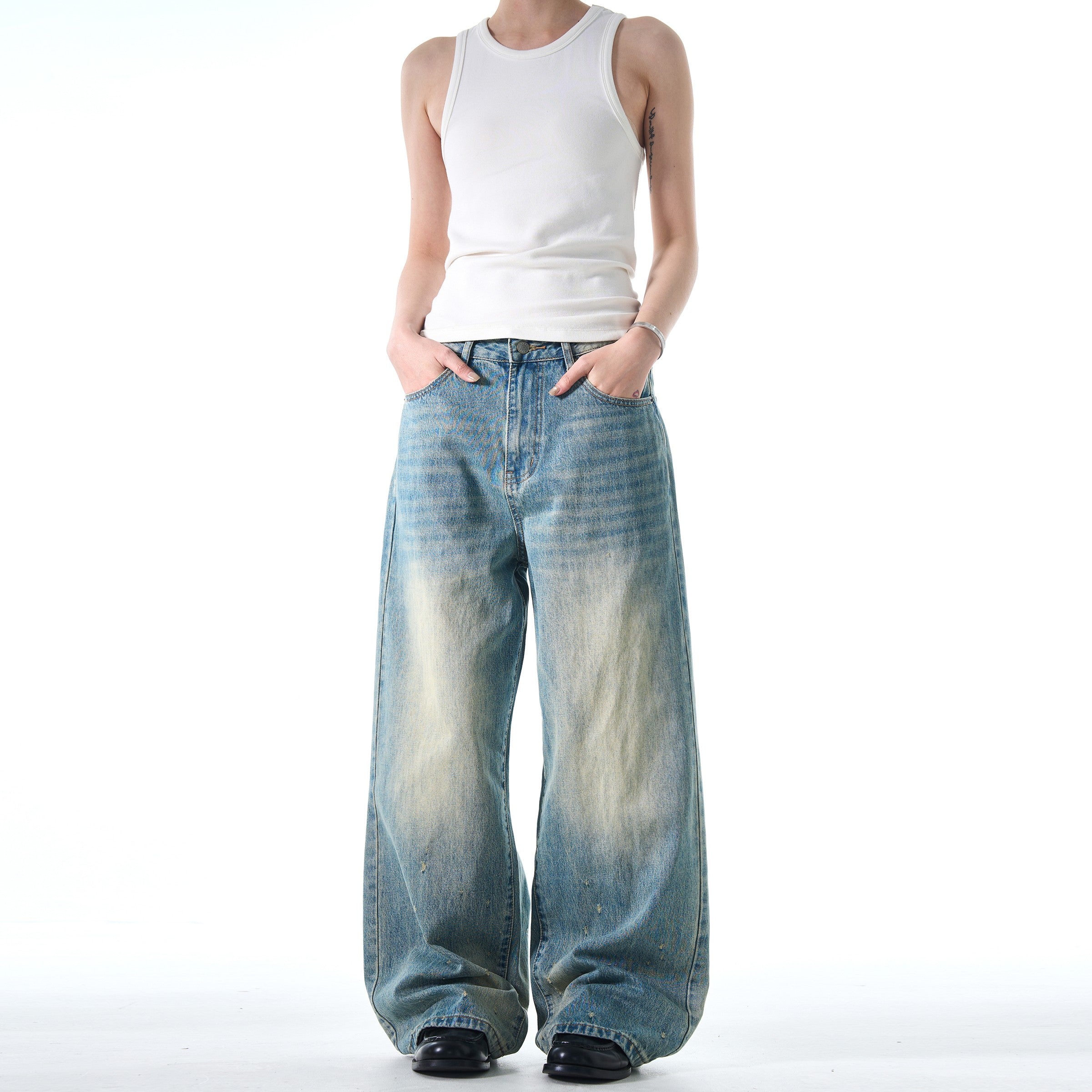 A person wearing a simple white tank top and black shoes stands with their hands in their pockets, modeling a pair of Retro Washed Jeans.