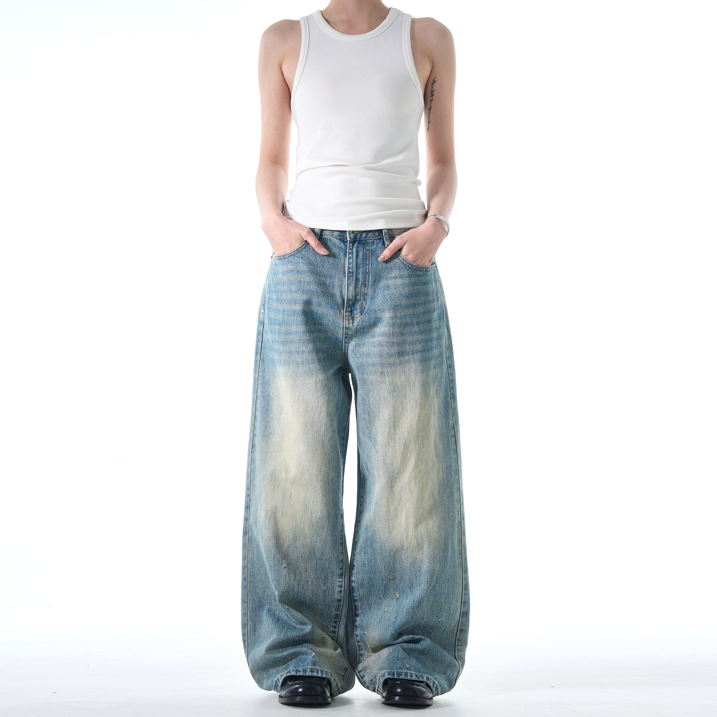 A person stands with hands in their pockets, wearing a white tank top and black shoes with a pair of loose, wide-legged Retro Jeans.