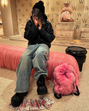 A person wearing a black beanie, a black leather jacket, and distressed jeans with rhinestones, sits on a long pink furry bench next to a matching pink bunny Plush Backpacks - Lost Series and scattered money, in a modern interior with "W" logos.