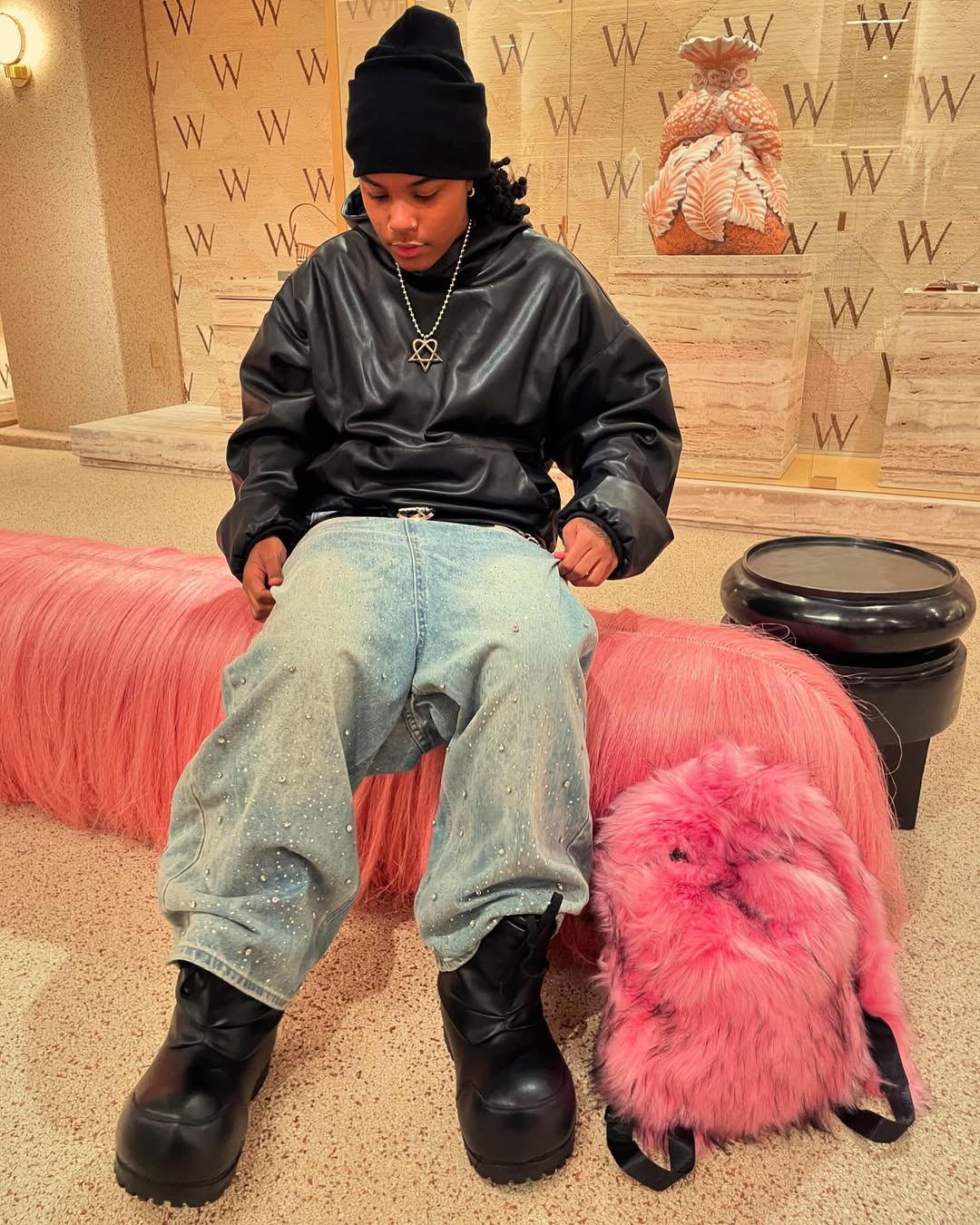 A person wearing a black leather jacket, and distressed jeans with rhinestones, sits on a long pink furry bench next to a matching pink bunny Plush Backpacks - Lost Series.