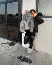 A person in a black jacket, leather pants, and fuzzy grey boots holds a matching grey Plush Backpacks - Lost Series over their face, standing beside a building with scattered items on the sidewalk.