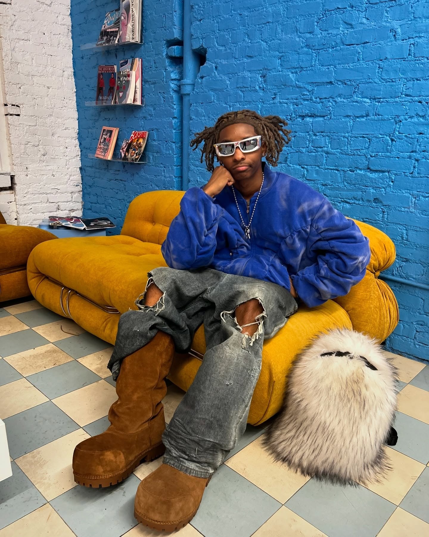 A person with dreadlocks and sunglasses, wearing a blue jacket, ripped jeans, and brown boots, sits on a mustard yellow couch next to a fluffy white Plush Backpacks - Lost Series, in a room with a blue brick wall.