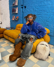 A person with dreadlocks and sunglasses, wearing a blue jacket, ripped jeans, and brown boots, sits on a mustard yellow couch next to a fluffy white Plush Backpacks - Lost Series, in a room with a blue brick wall.