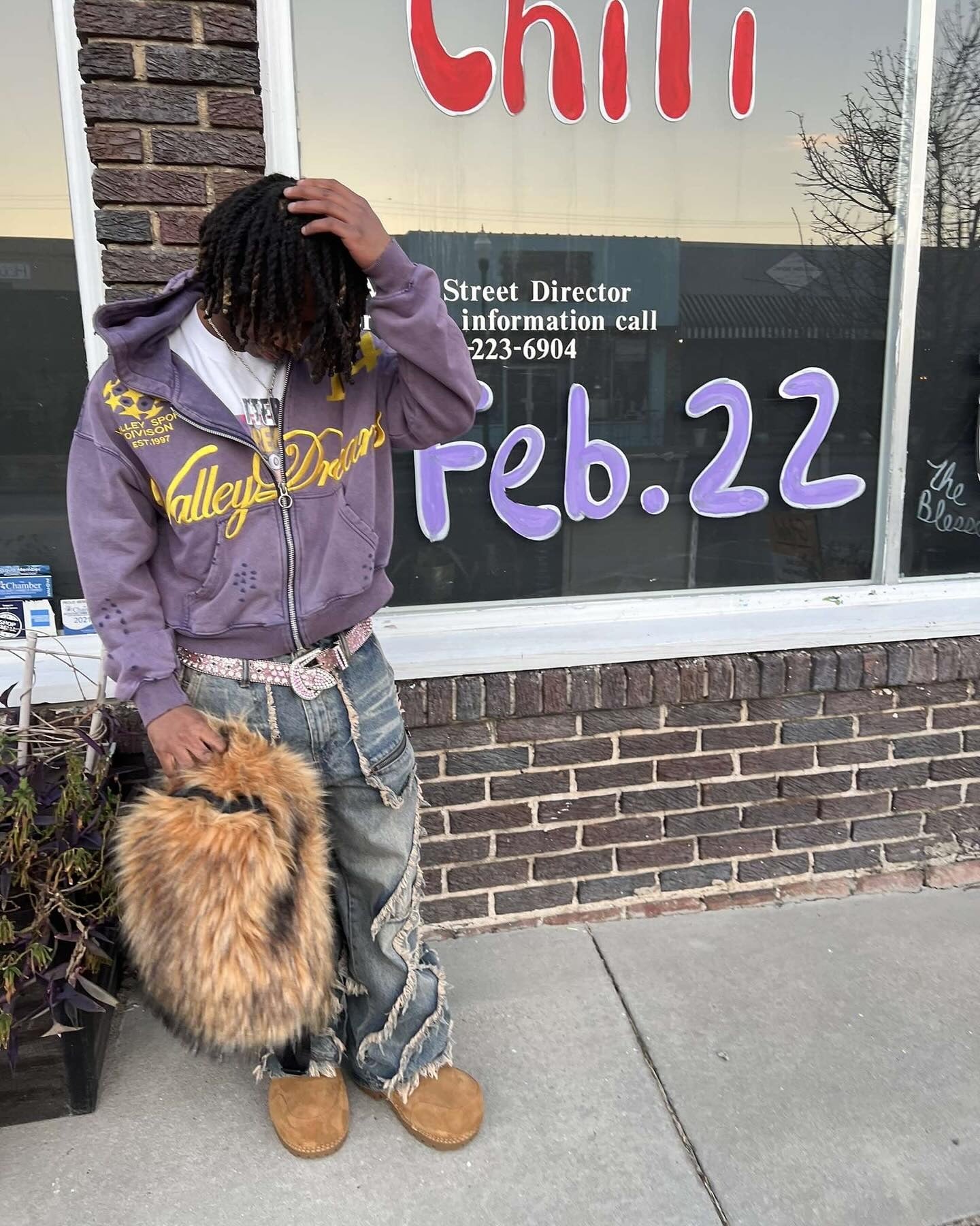 A person with dreadlocks wears a purple graphic hoodie, distressed jeans, and tan shoes, holding a fluffy brown Plush Backpacks - Lost Series while posing in front of a storefront with "Feb. 22" painted on the window.
