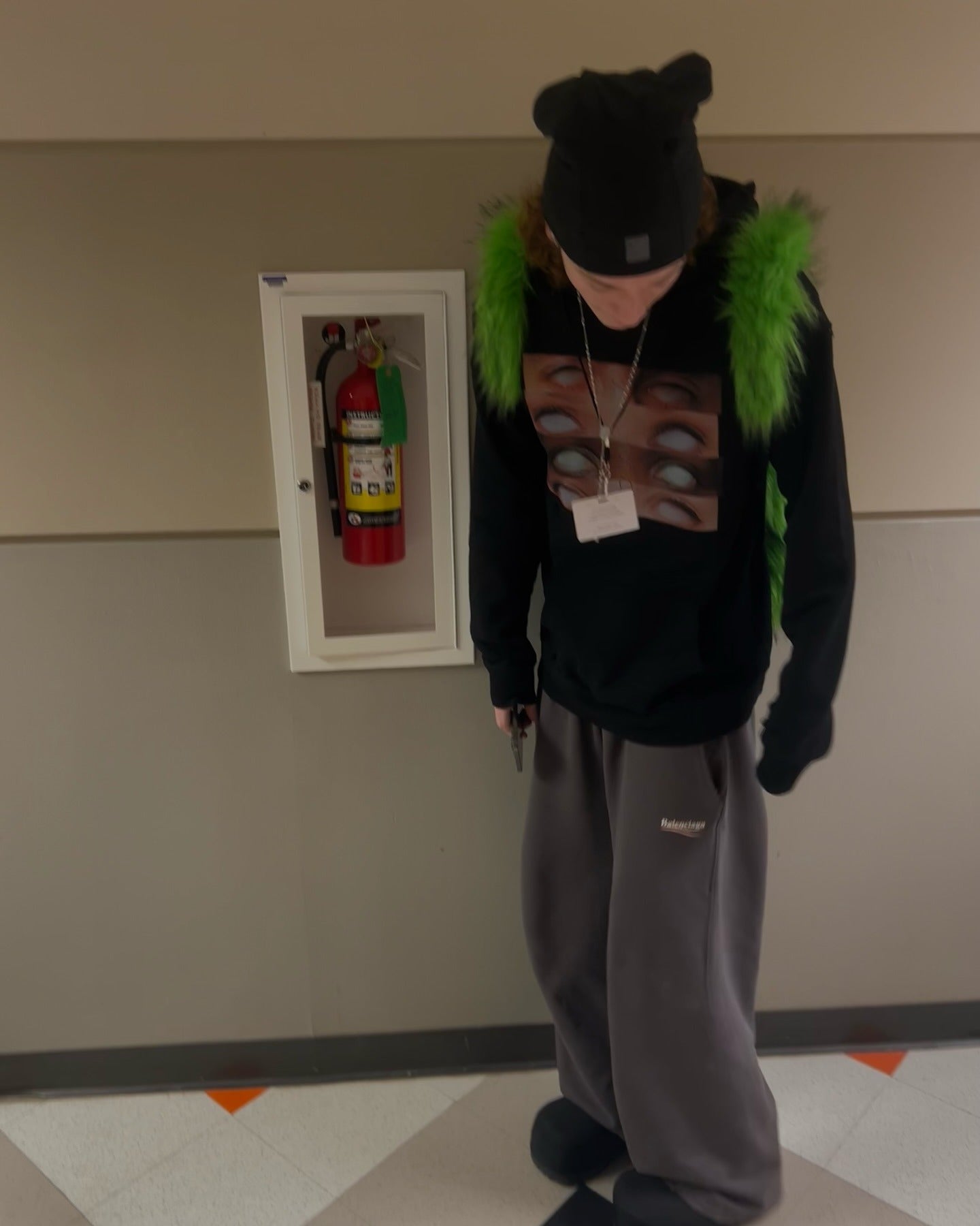 A person wearing a black beanie, a black hoodie with an eye graphic, grey sweatpants, and black shoes, stands indoors with a neon green furry Plush Backpacks - Lost Series visible on their shoulders, next to a fire extinguisher cabinet.