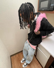 A person with long dreadlocks, wearing a black zip-up hoodie with a pink fluffy trim and light-wash baggy jeans with a matching pink belt, leans on a kitchen counter, showcasing a pink Plush Backpacks - Lost Series or a similar accessory.