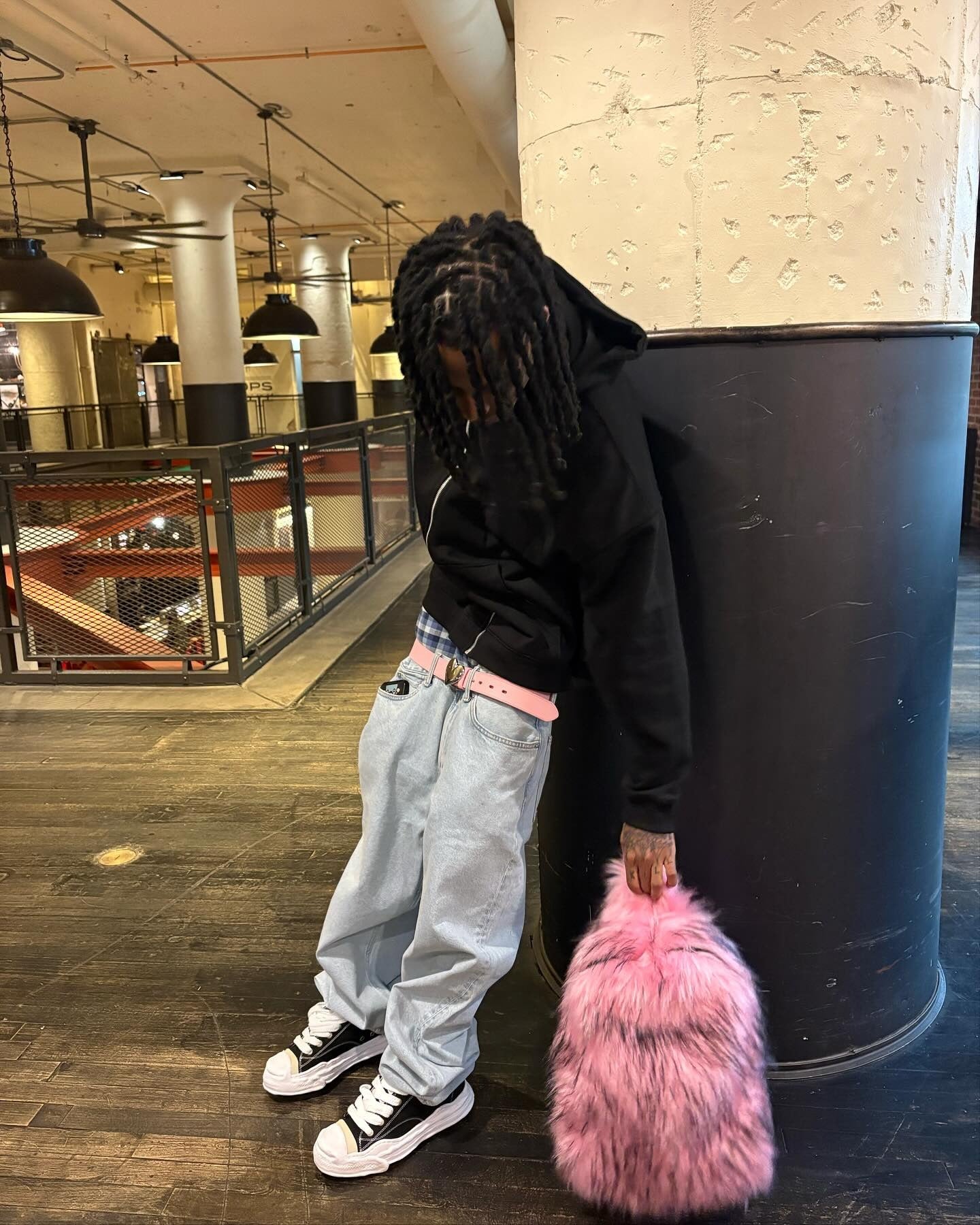 A person with dreadlocks, wearing a black hoodie, light-wash baggy jeans, and black sneakers, leans against a large pillar while holding a fluffy hot pink and black Plush Backpacks - Lost Series.