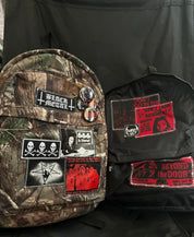 A brown camouflage backpack with various black and white patches and some pins sits next to a black PT269 Dead Control Backpack featuring red and black horror-themed patches.