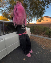 A person wearing a black graphic t-shirt, baggy black pants with white stitching, and pink slippers leans against a white car while holding a fluffy pink Plush Backpacks - Lost Series over their head, showcasing a playful and vibrant design.