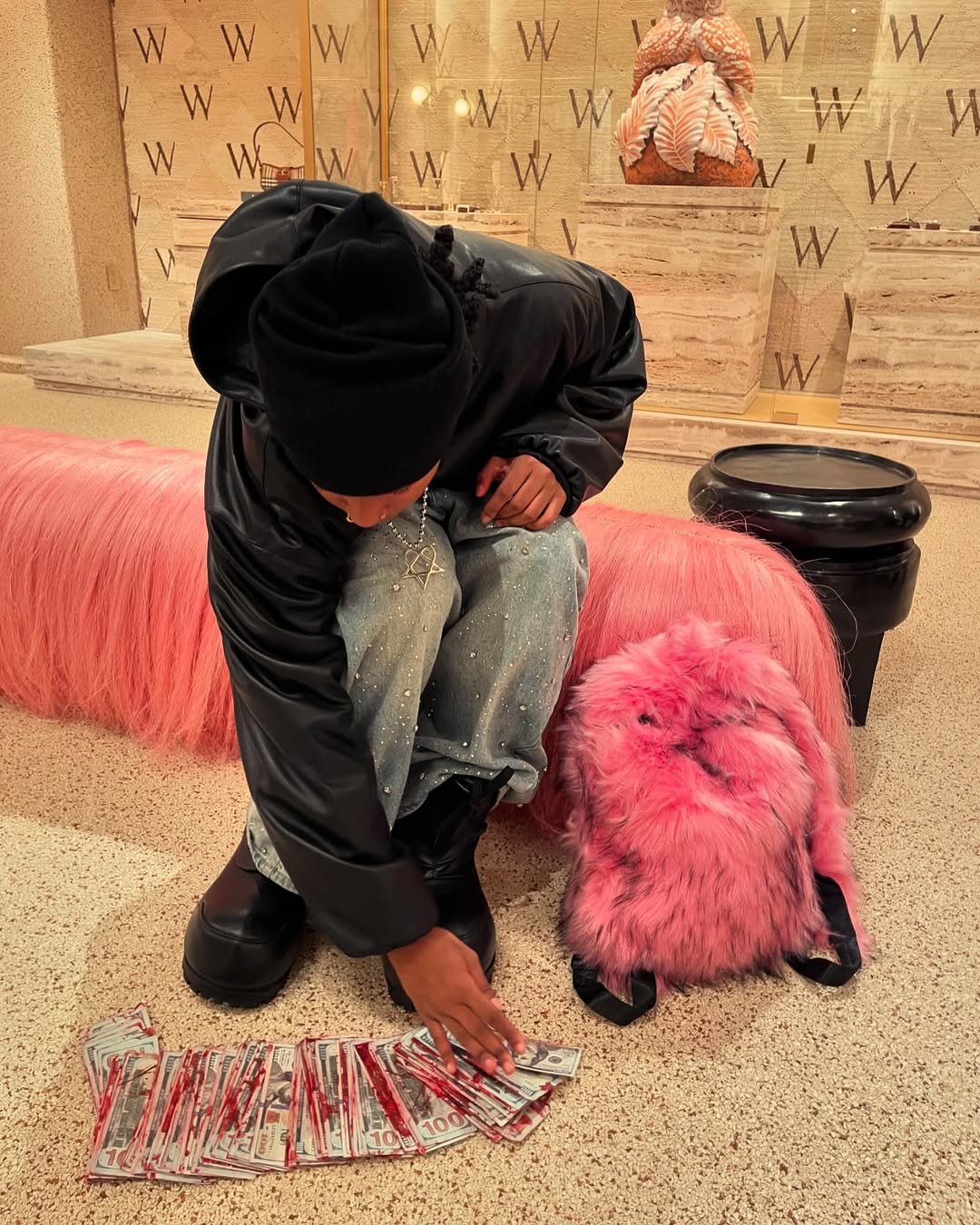 A person wearing a black beanie, a black leather jacket, and distressed jeans with rhinestones, sits on a long pink furry bench next to a matching pink bunny Plush Backpacks - Lost Series and scattered money, in a modern interior with "W" logos.