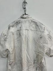 Back of a white men's graffiti t-shirt with short sleeves, featuring an all-over gray and black distressed or painted effect, hanging on a white wall.