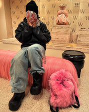 A person in a black beanie, leather jacket, and rhinestone-studded jeans sits on a pink furry bench, holding fanned money to their face, with a matching pink Plush Backpacks - Lost Series beside them in a "W" branded room.