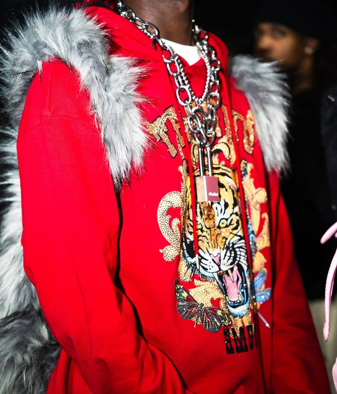 A person wears a vibrant red hoodie with a detailed tiger and ornate gold embroidery, layered with multiple chunky silver chains and a grey furry Plush Backpacks - Lost Series or similar accessory on their back.