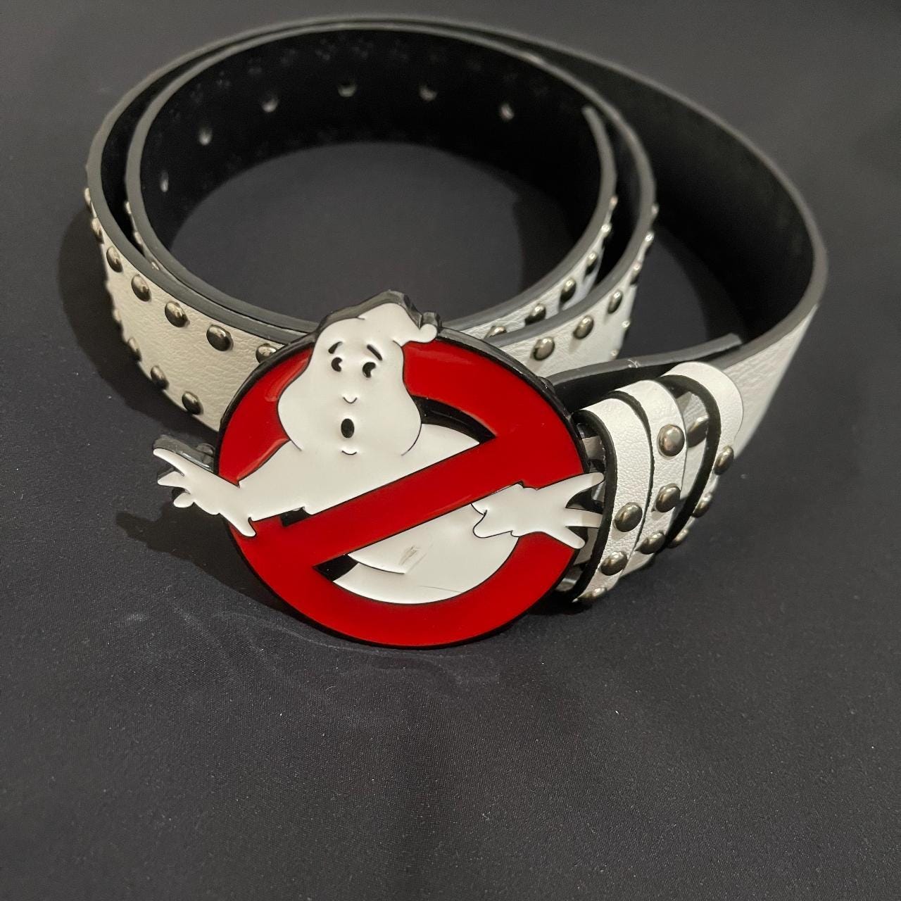 Ghost Buster Belt with silver studs is shown on a black surface, highlighting the large Ghostbusters logo buckle with the iconic ghost figure in a red circle.