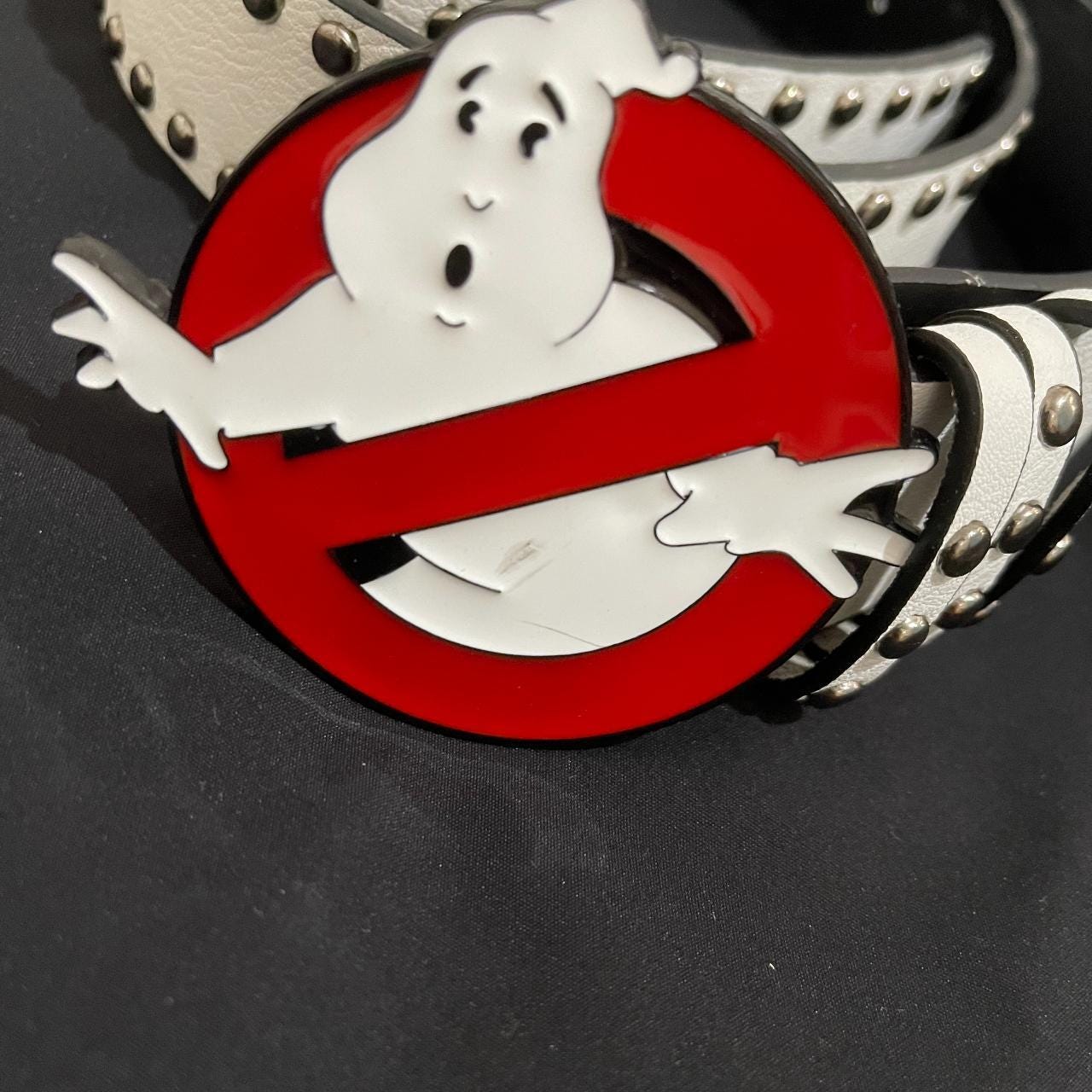A close-up of a Ghost Busters Belt shows a large, round buckle with the classic ghost-in-a-red-circle logo on a white studded belt against a black background.