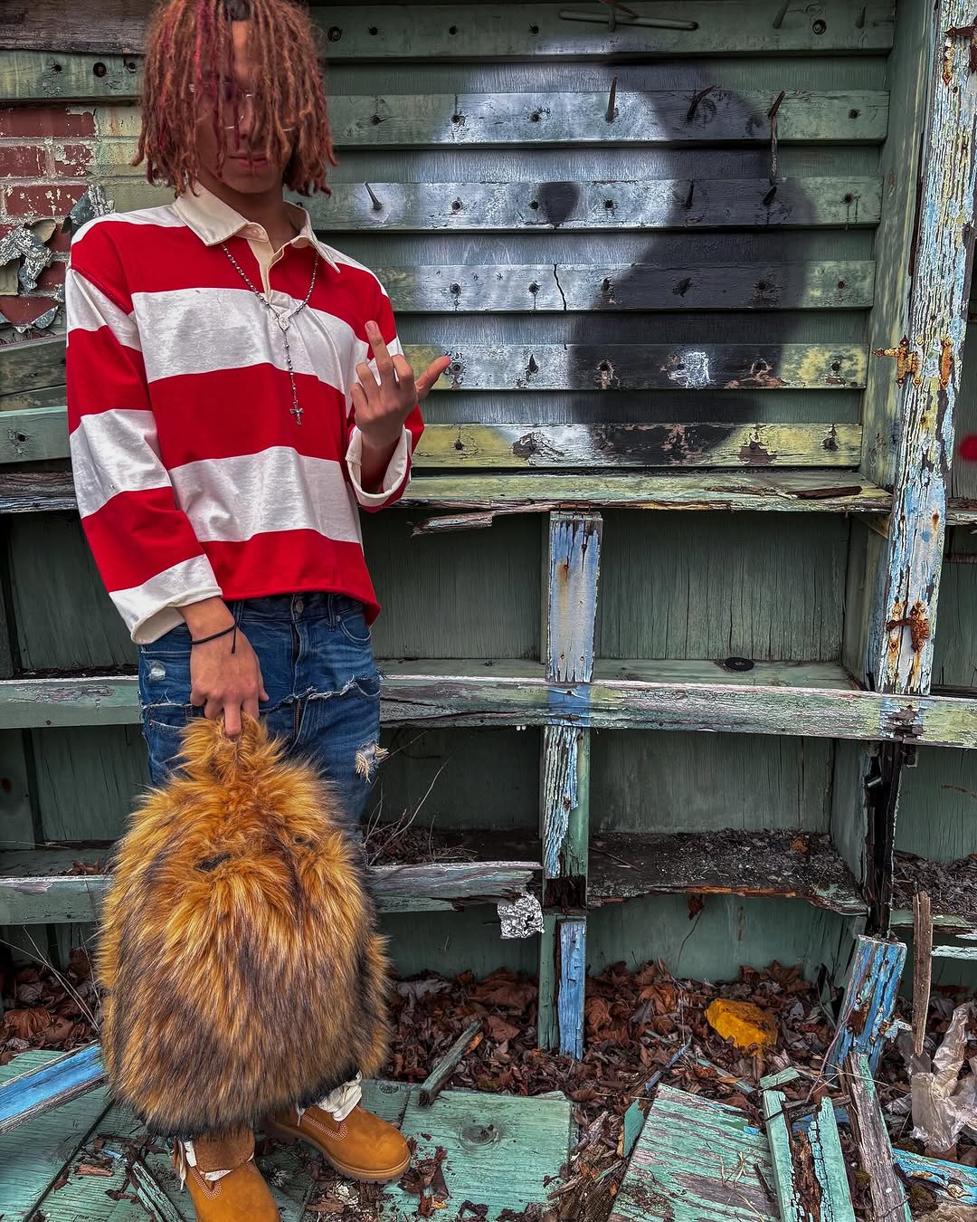 A person with red dreadlocks, wearing a red and white striped polo shirt and distressed jeans, holds a brown fluffy Plush Backpacks - Lost Series while standing in front of a weathered wooden structure.