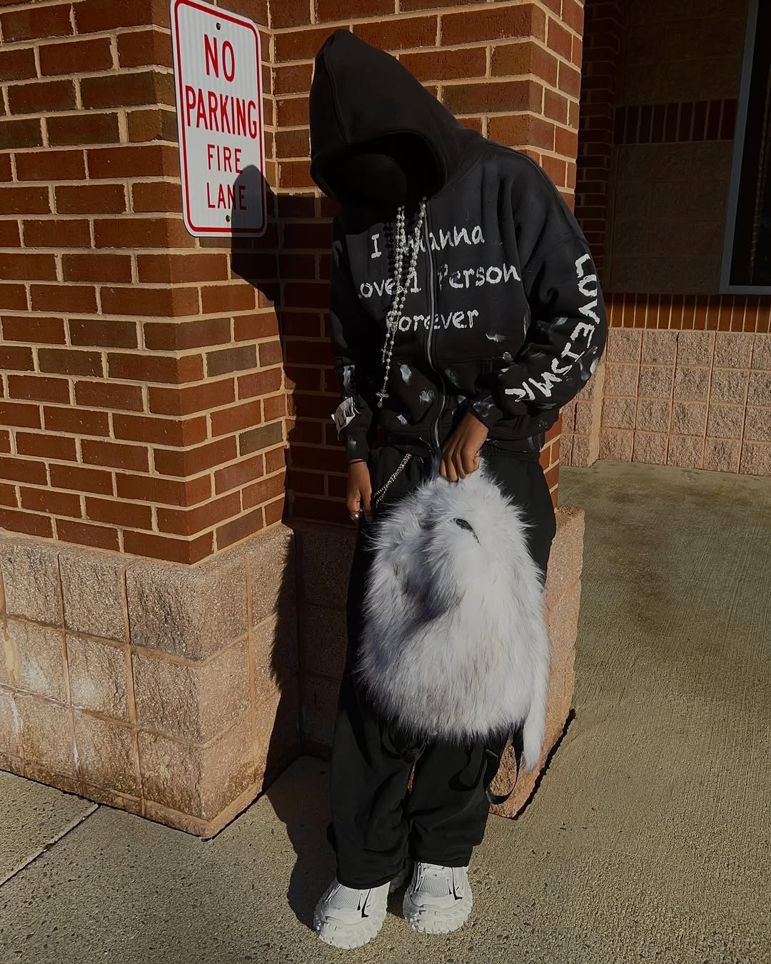 A person wearing a black hoodie and distressed jeans holds a fluffy white Plush Backpacks - Lost Series over their shoulder while standing in front of a brick building.