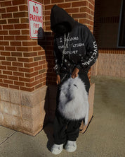 A person wearing a black hoodie and distressed jeans holds a fluffy white Plush Backpacks - Lost Series over their shoulder while standing in front of a brick building.