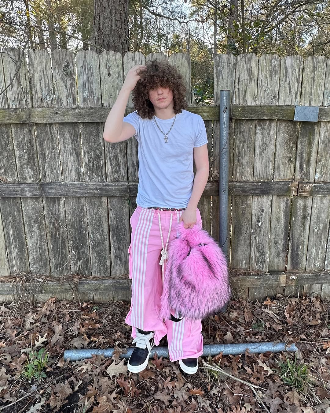 A person with curly hair, wearing a white t-shirt, pink striped track pants, and black sneakers, stands in front of a wooden fence while holding a fluffy hot pink and black Plush Backpacks - Lost Series.