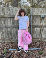 A person with curly hair, wearing a white t-shirt, pink striped track pants, and black sneakers, stands in front of a wooden fence while holding a fluffy hot pink and black Plush Backpacks - Lost Series.