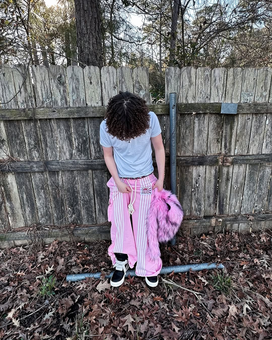 A person with curly hair,wearing white t-shirt and pink striped pants holds a hot pink Plush Backpacks - Lost Series in front of a wooden fence.