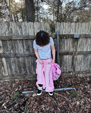 A person with curly hair,wearing white t-shirt and pink striped pants holds a hot pink Plush Backpacks - Lost Series in front of a wooden fence.