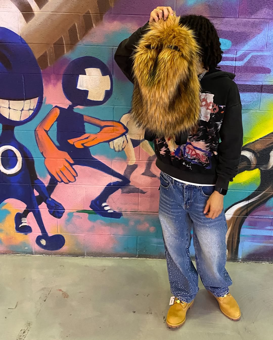 A person wearing a black graphic hoodie, distressed jeans, and tan boots holds a fluffy brown Plush Backpacks - Lost Series over their head, standing in front of a colorful graffiti wall with cartoon characters.