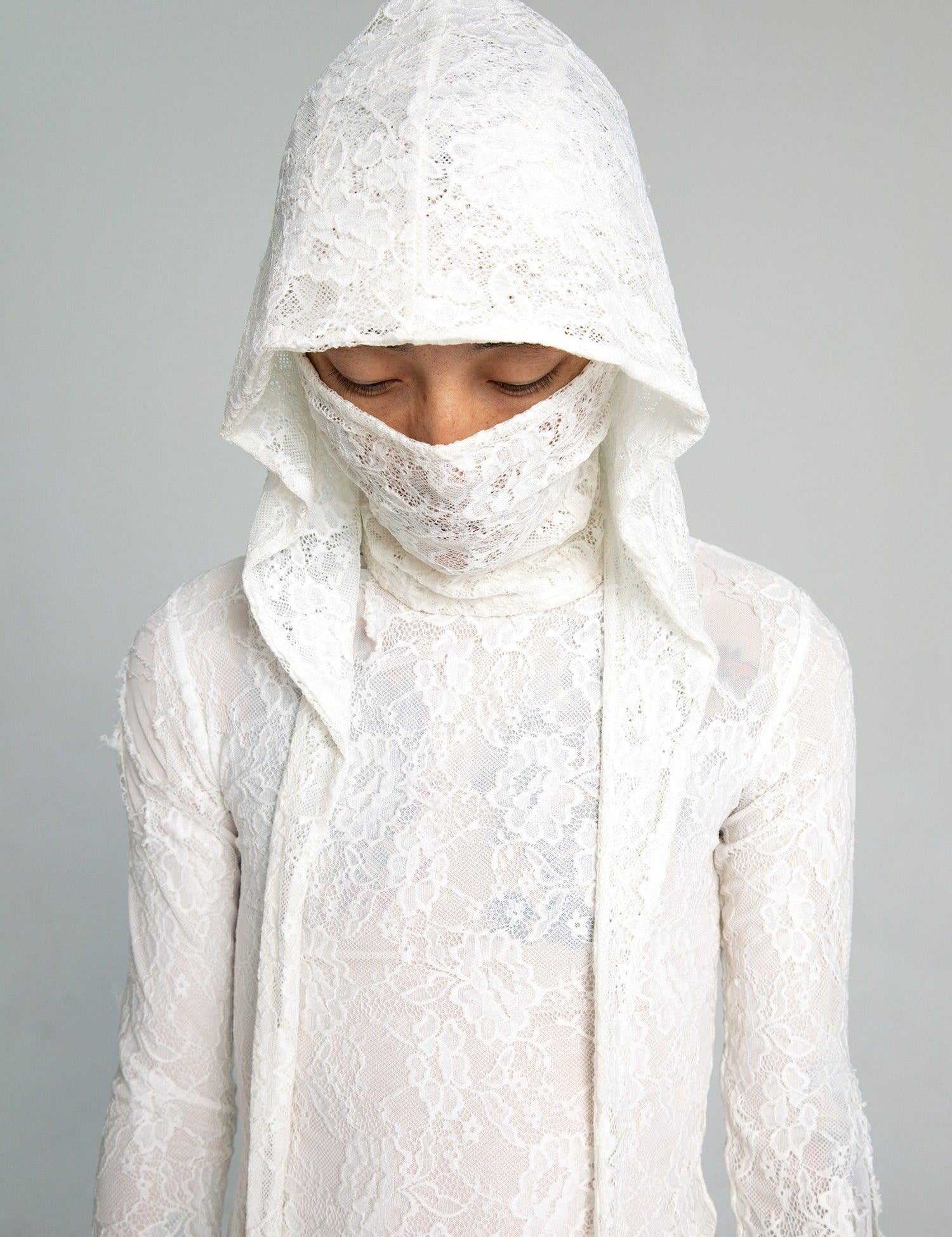 A person wears a semi-sheer white lace top with a lace hood mask that covers the face, with their eyes closed and head slightly bowed.