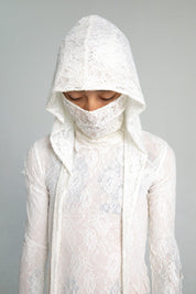 A person wears a semi-sheer white lace top with a lace hood mask that covers the face, with their eyes closed and head slightly bowed.