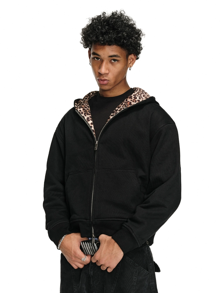 A person with curly dark hair wears a black Leopard Lined Jacket with the zipper partially open, revealing a black shirt underneath, and holds their striped belt with both hands.