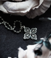 A close-up of a silver NightMelody GothRite Necklace with an ornate floret-shaped pendant resting on a dark, textured surface next to a soft, white fabric.