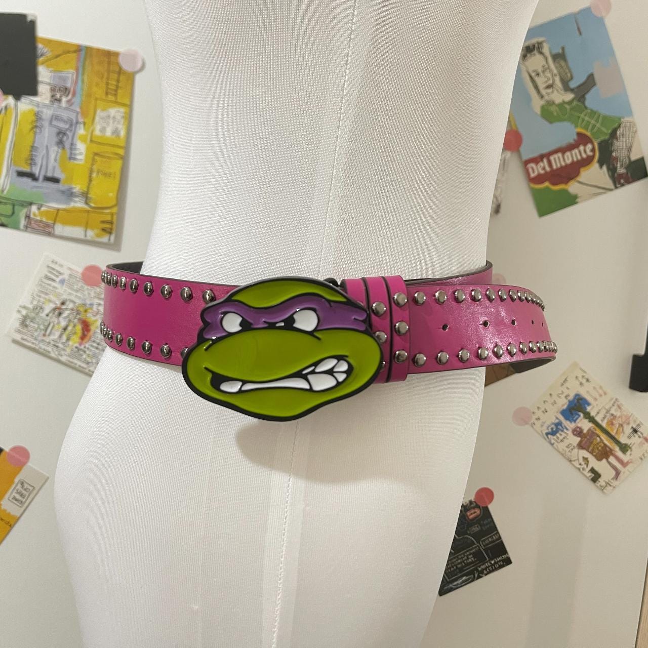 A pink studded ninja turtle belt with a large Donatello face as the buckle is shown worn on a white mannequin against a background of various posters and artwork.