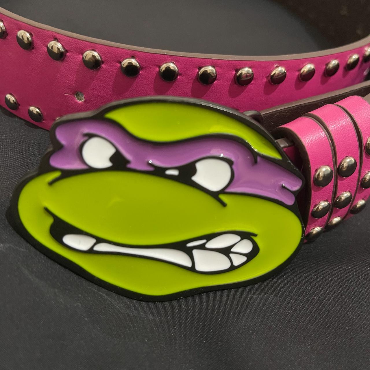 A close-up shot highlights a vibrant green Donatello ninja turtle buckle with a purple mask, attached to a studded pink belt.