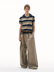 A woman posing in a polo shirt retro with horizontal navy and tan stripes. She is also wearing loose-fitting tan pants with several layered belts and has her hands in her pockets.