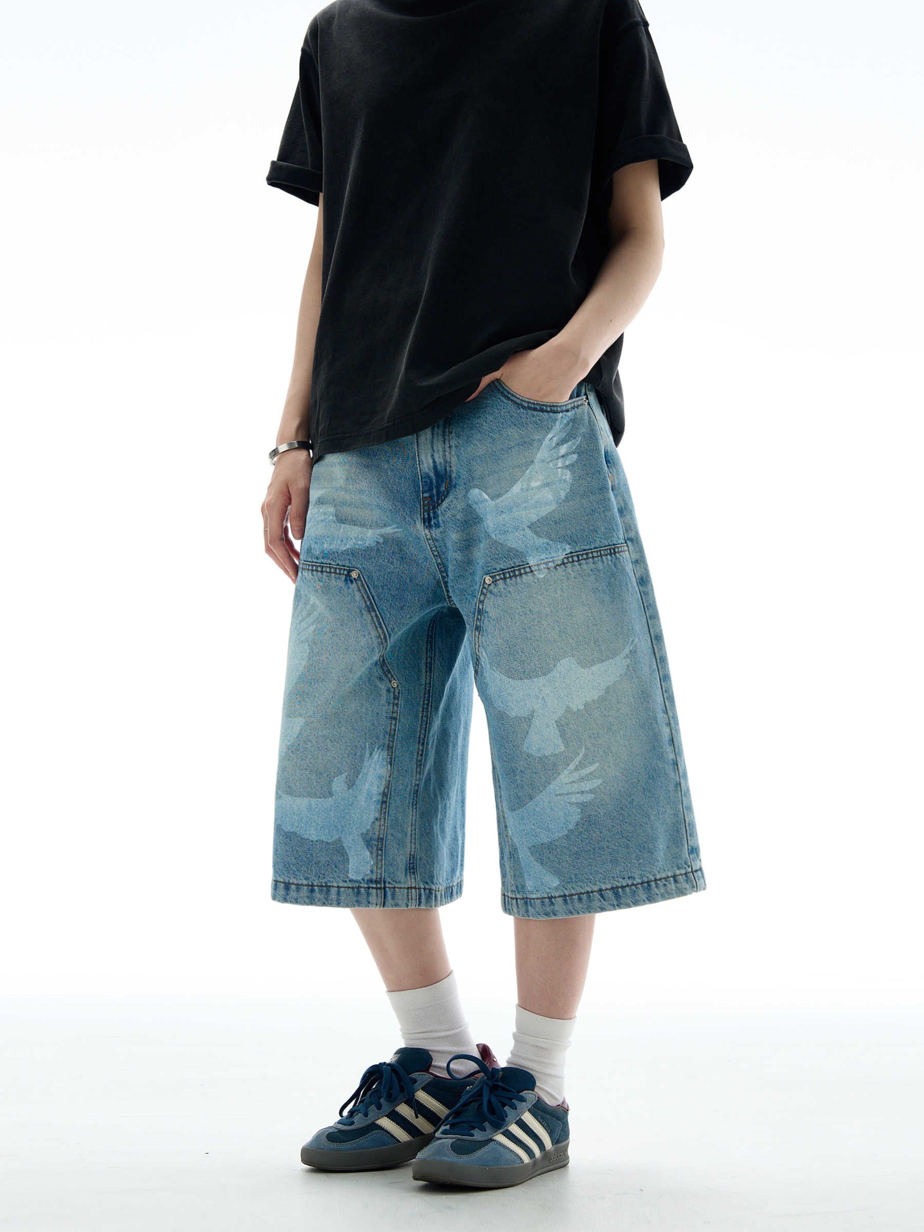 A person stands wearing a black t-shirt and light blue SkyBound Shorts with faded bird silhouettes, white socks, and blue and white sneakers.