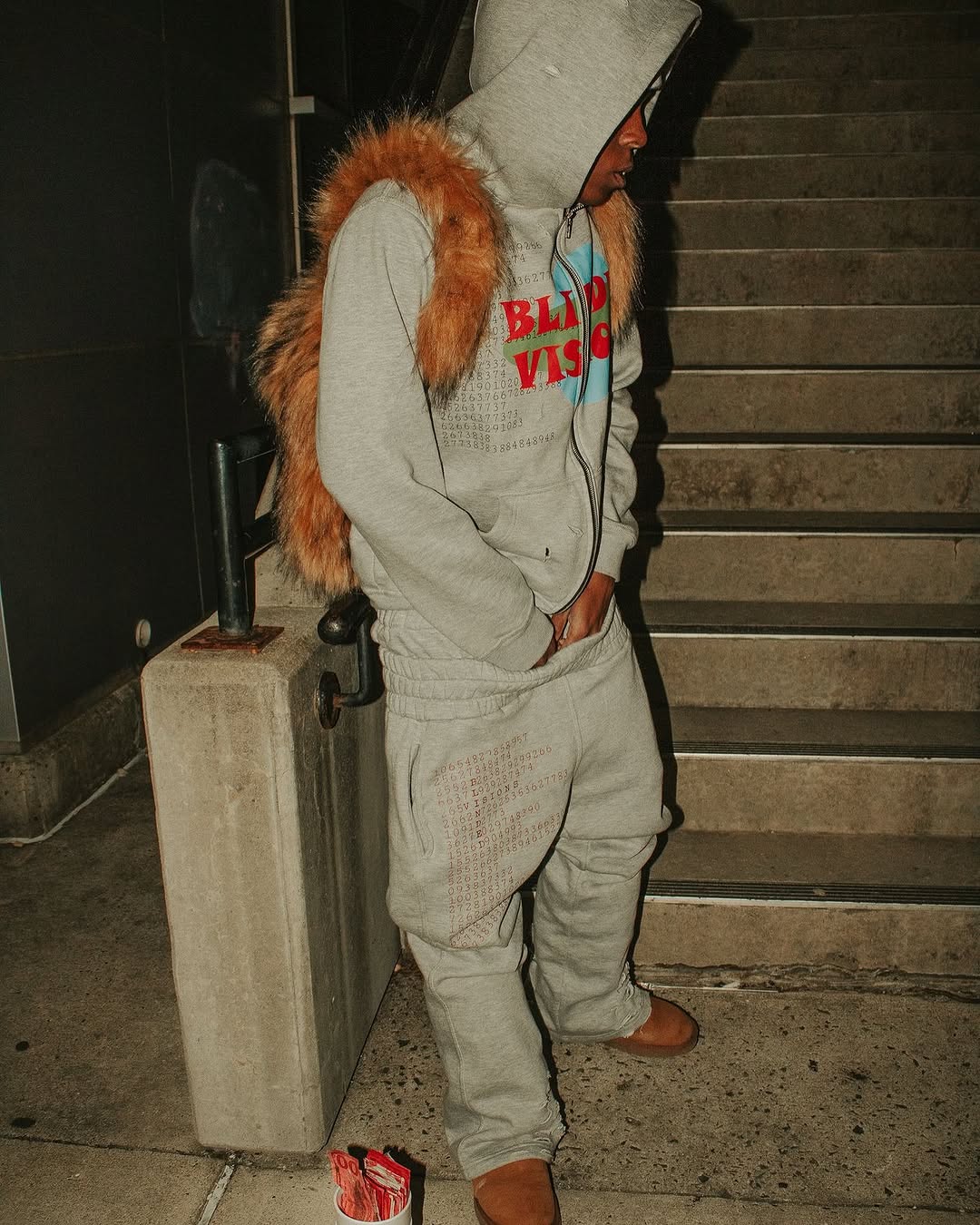 A person wearing a grey hooded sweatshirt with colorful text, matching grey sweatpants, and brown shoes stands by concrete stairs, with a fluffy brown Plush Backpacks - Lost Series draped over their shoulders.