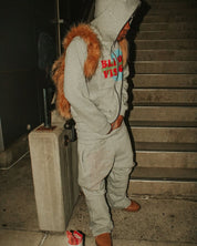 A person wearing a grey hooded sweatshirt with colorful text, matching grey sweatpants, and brown shoes stands by concrete stairs, with a fluffy brown Plush Backpacks - Lost Series draped over their shoulders.