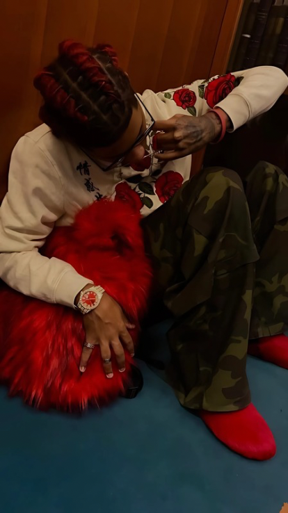 A person with braided red hair, wearing a white rose-embroidered hoodie and camouflage pants, sits on the floor while hugging a fluffy red Plush Backpacks - Lost Series.