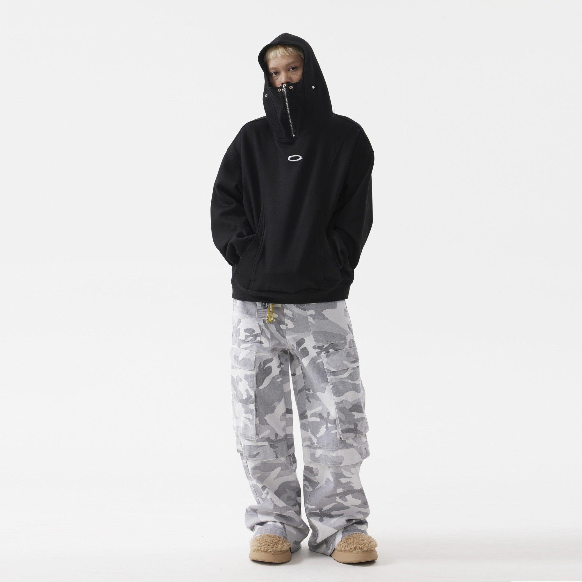 A person with blonde hair stands with their hands in their pockets, wearing a black hoodie with mask and light-colored camouflage pants.