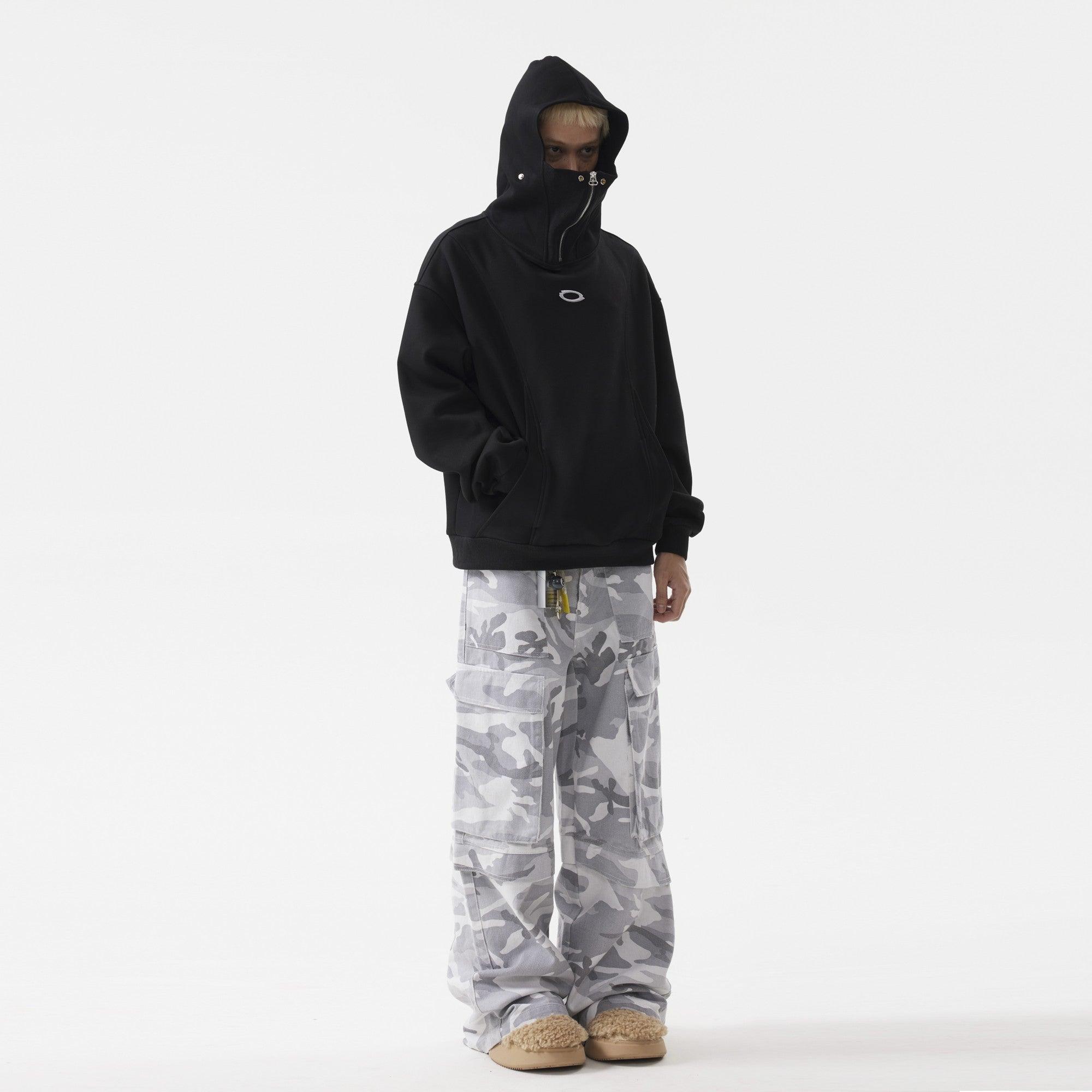 A man with black masked hoodie with a zipper, paired with gray camouflage cargo pants and tan slippers, standing against a white background.