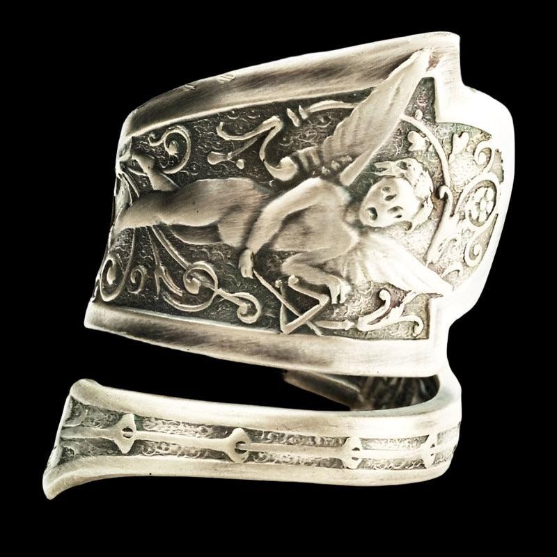 A silver fallen angel ring with an ornate, engraved design of a cherubic figure and musical notes is shown on a solid black background.