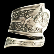 A silver fallen angel ring with an ornate, engraved design of a cherubic figure and musical notes is shown on a solid black background.