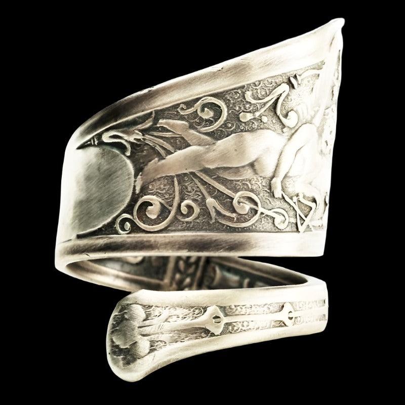 Silver fallen angel ring with a detailed depiction of a winged figure and intricate patterns is displayed on a dark surface.