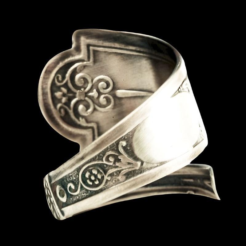 A close-up of a sculpted silver fallen angel ring featuring an embossed cherub and musical motifs, set against a black backdrop.