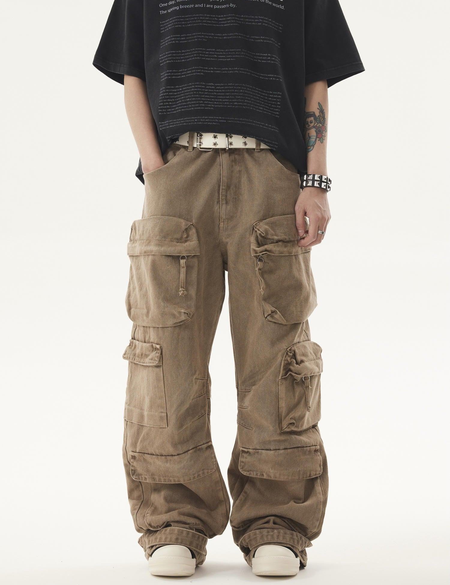 A person stands wearing a black t-shirt and Heavy duty Vibe Cargo Jeans with multiple large pockets, accessorized with a white belt and studded bracelet, with navigation arrows on the sides of the image.