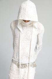 A person with their head bowed wears a semi-sheer white lace top with a matching lace mask hood that covers their face, with a wide studded belt and white distressed pants.