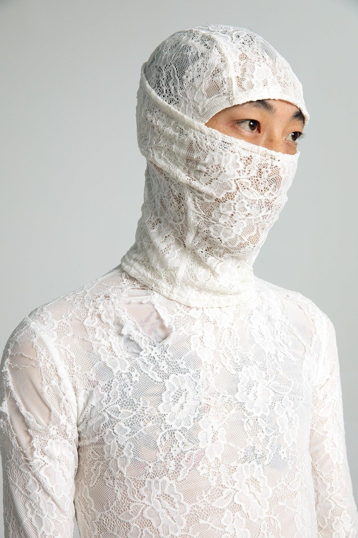 A man wears a semi-sheer white lace top with a lace mask hood that covers most of their face, looking to the side with their eyes visible.