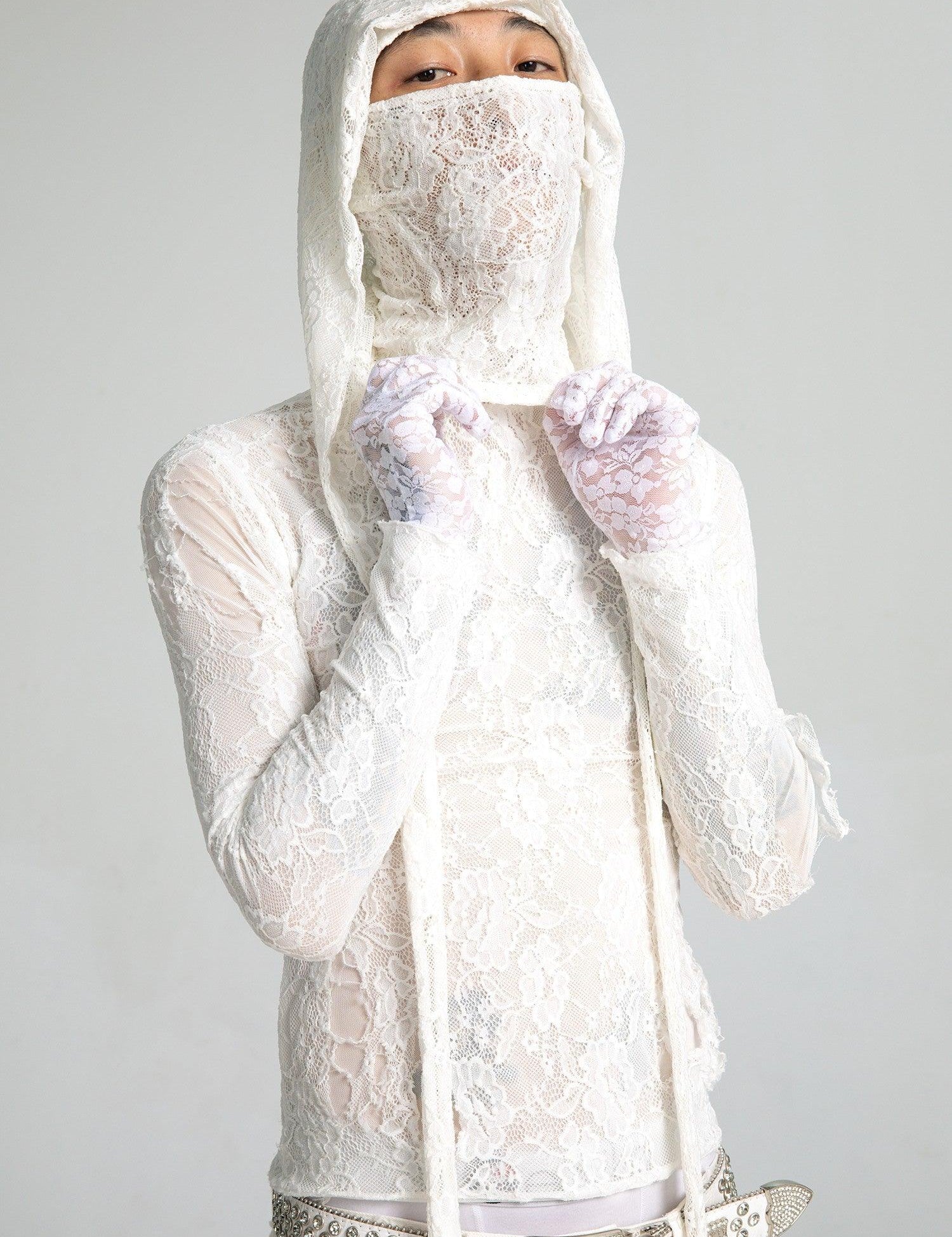 A person looks directly at the camera, wearing a semi-sheer white lace top with a matching lace hood mask and lace gloves