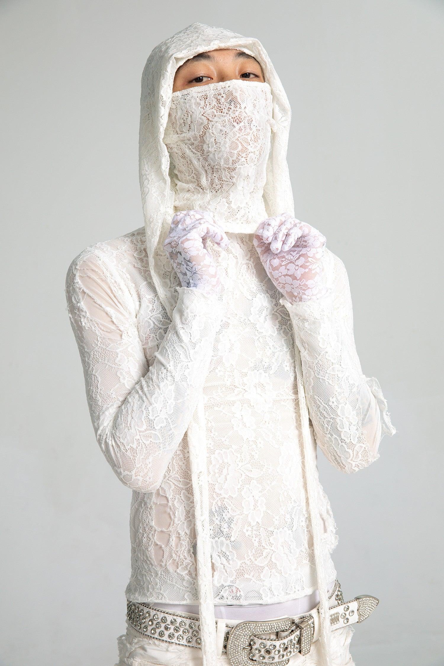 A person looks directly at the camera, wearing a semi-sheer white lace top with a matching lace hood mask and lace gloves